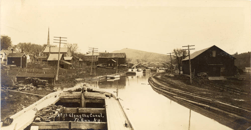 crandall_1660_large Canal boat in foreground on the canal with houses, barns, a church, and utility poles on either side, with another boat along the edge and an iron bridge in the background.